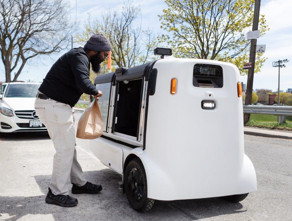 A person loading a delivery bag into a white three-wheeled autonomous delivery robot on a city street.